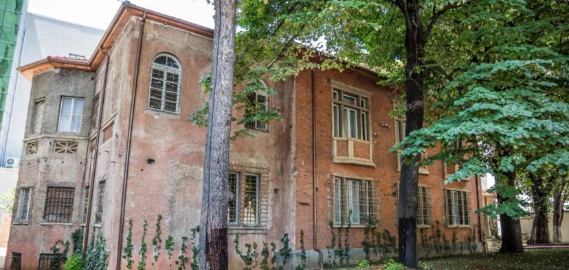 Exterior view of a historic building in Tirana, Albania, showcasing a mix of brick and plaster textures, surrounded by lush greenery.